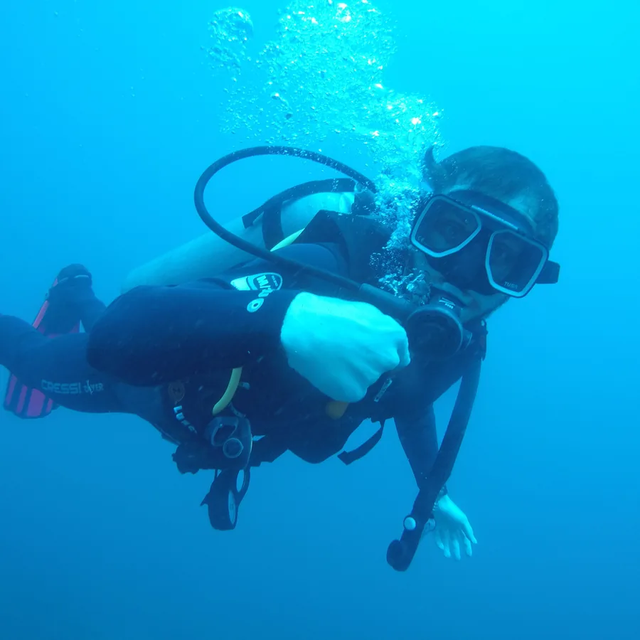 Tomer scuba diving in clear blue water, holding an underwater camera, a stream of bubbles rising from his regulator.