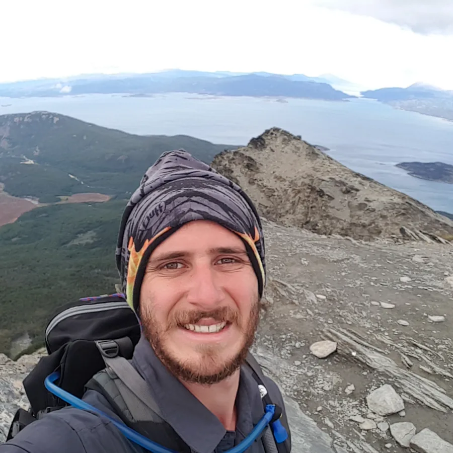 Tomer on a rocky summit ridge in a beanie and backpack, with forested mountains and a wide island-scattered channel far below.